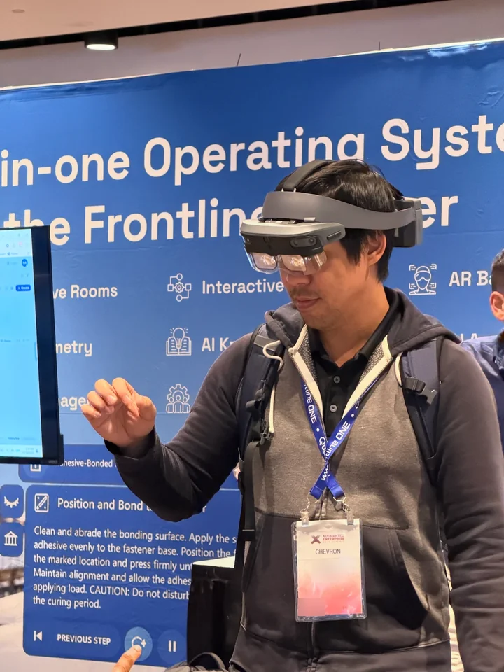 An attendee wears an AR headset in front of a blue booth backdrop about an operating system for the frontline worker