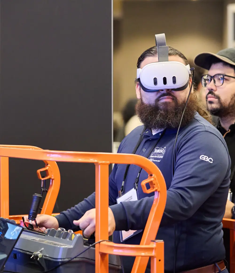 An attendee wearing a VR headset works the controls of an orange aerial work platform simulator as an onlooker watches