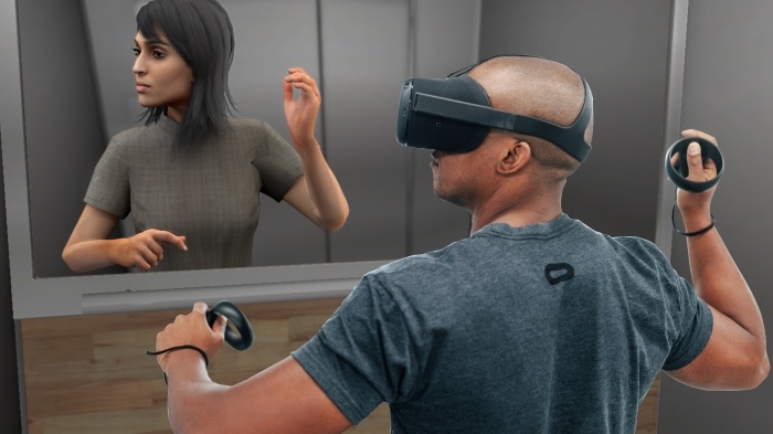A man wearing a virtual reality headset and controllers, interacting with a digital image of a woman in a mirrored environment.