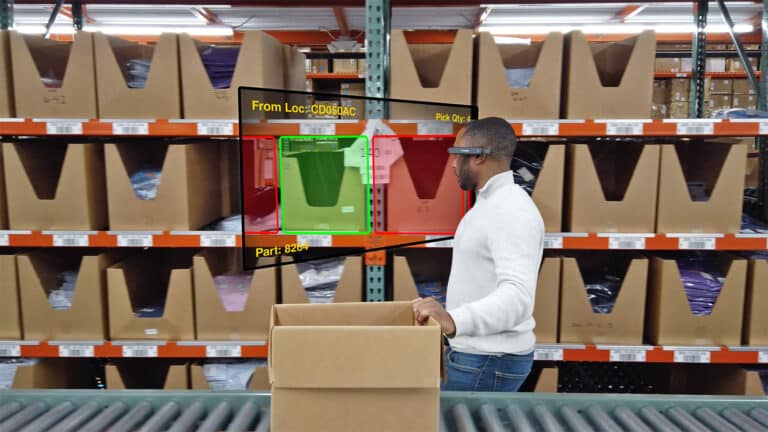 A man in a white sweater using a wearable computer device to scan a package in a warehouse with shelves of boxes
