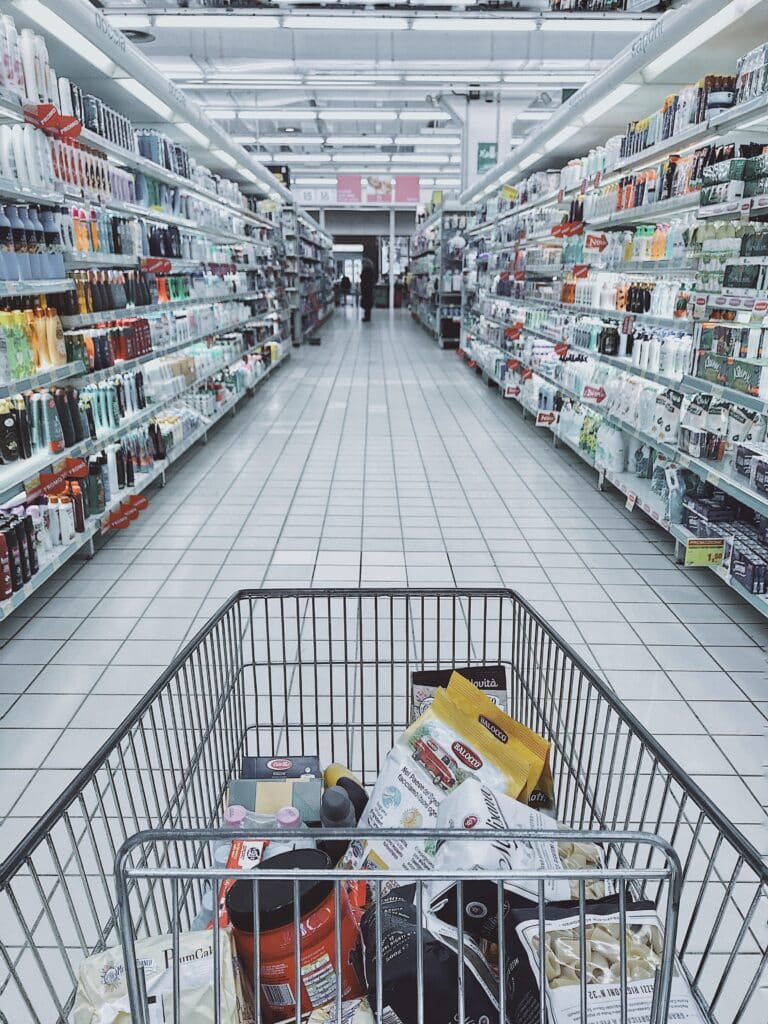 A shopping cart filled with various grocery items in the aisle of a grocery store, with shelves of products lining the walls on both sides.