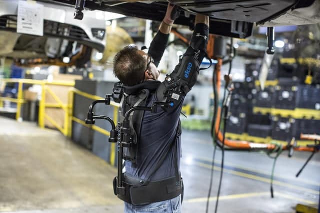A man wearing a harness and safety gear works on an overhead vehicle in an automotive manufacturing facility