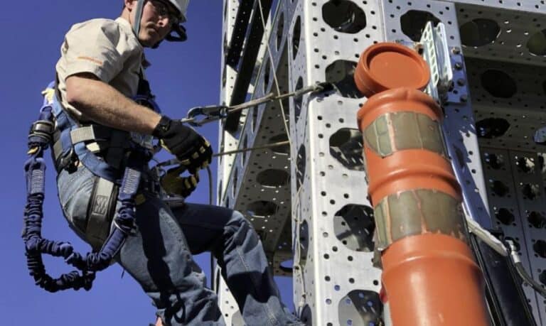 A worker in harness and protective gear climbing on industrial equipment against a blue sky