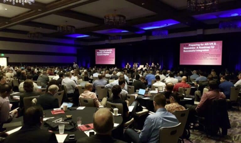 A large conference room filled with audience members listening to a presentation on a large screen at the front of the room.