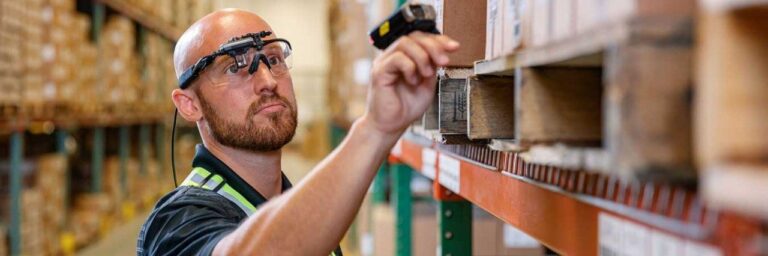 A construction worker wearing a hardhat and safety goggles inspects an industrial shelving unit in a warehouse.