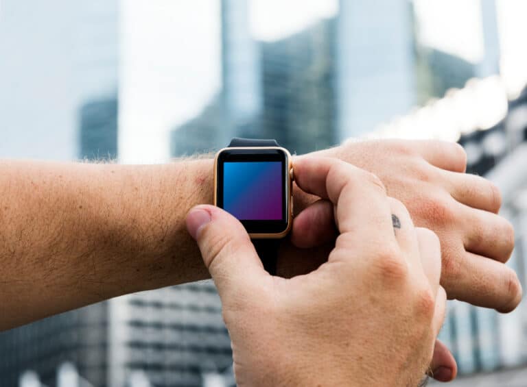 A person's hand wearing a smartwatch with a colorful display against a blurred cityscape background