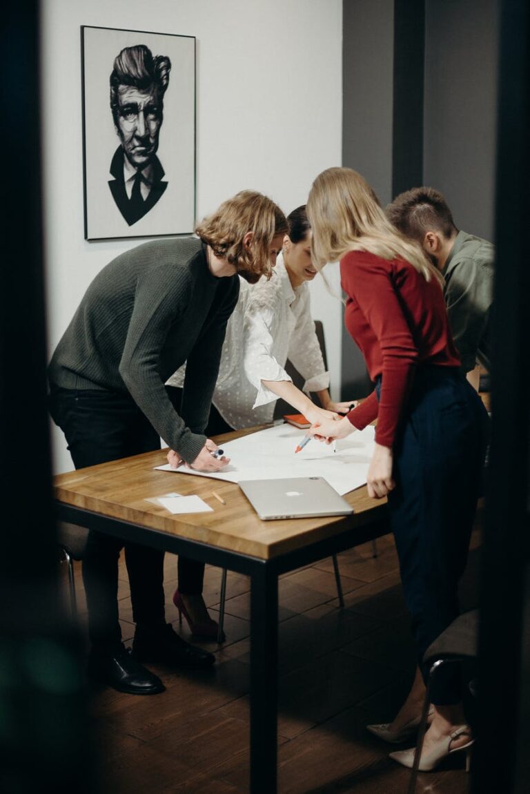 A group of people collaborating around a table in a creative office space, with a framed portrait of a man on the wall behind them.