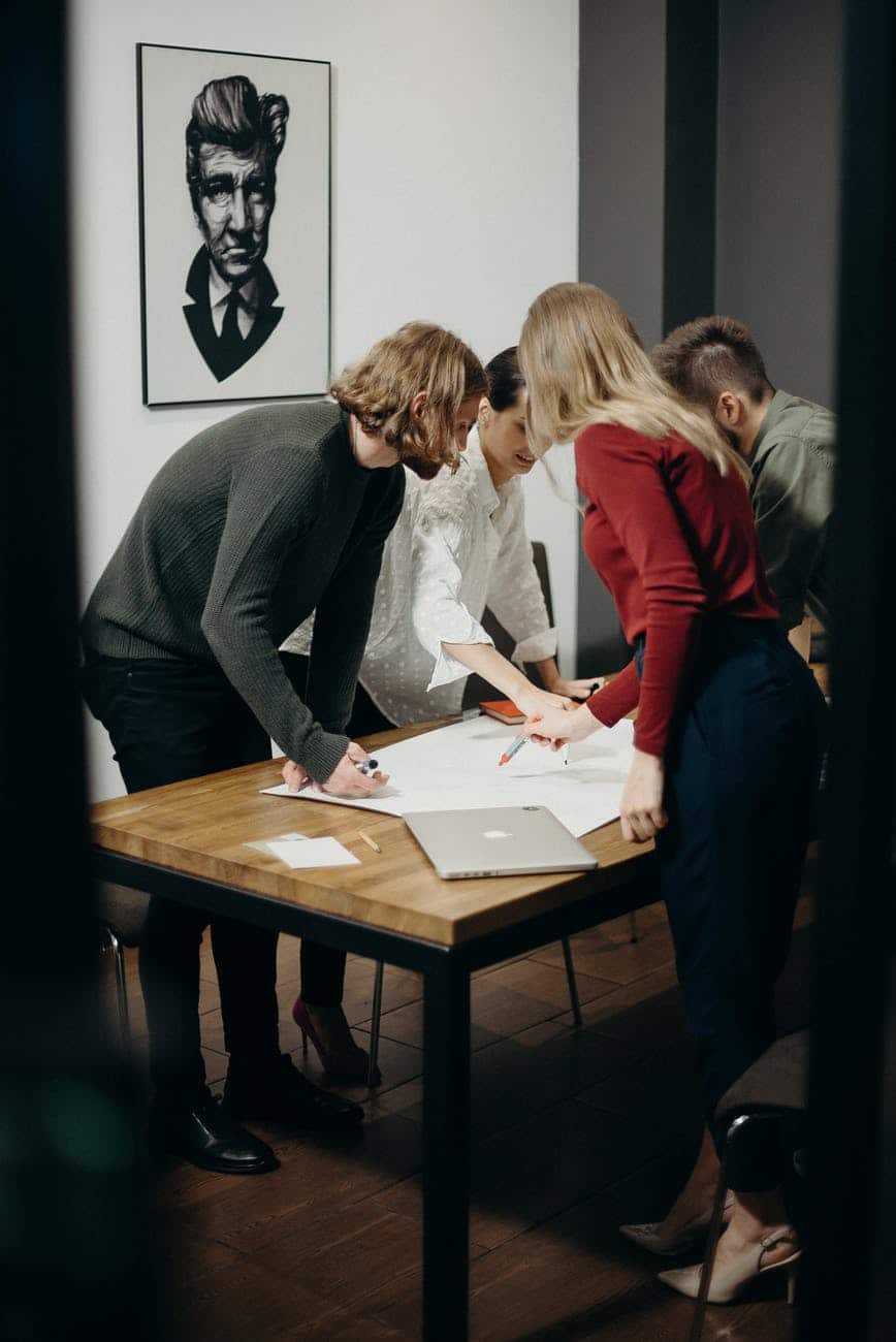 A group of people collaborating around a table in a creative office space, with a framed portrait of a man on the wall behind them.