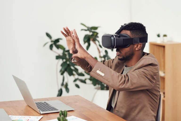 A man wearing a virtual reality headset gesturing with his hands in a home office setting with a laptop and plants in the background.