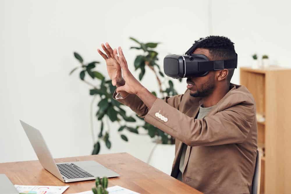 A man wearing a virtual reality headset gesturing with his hands in a home office setting with a laptop and plants in the background.