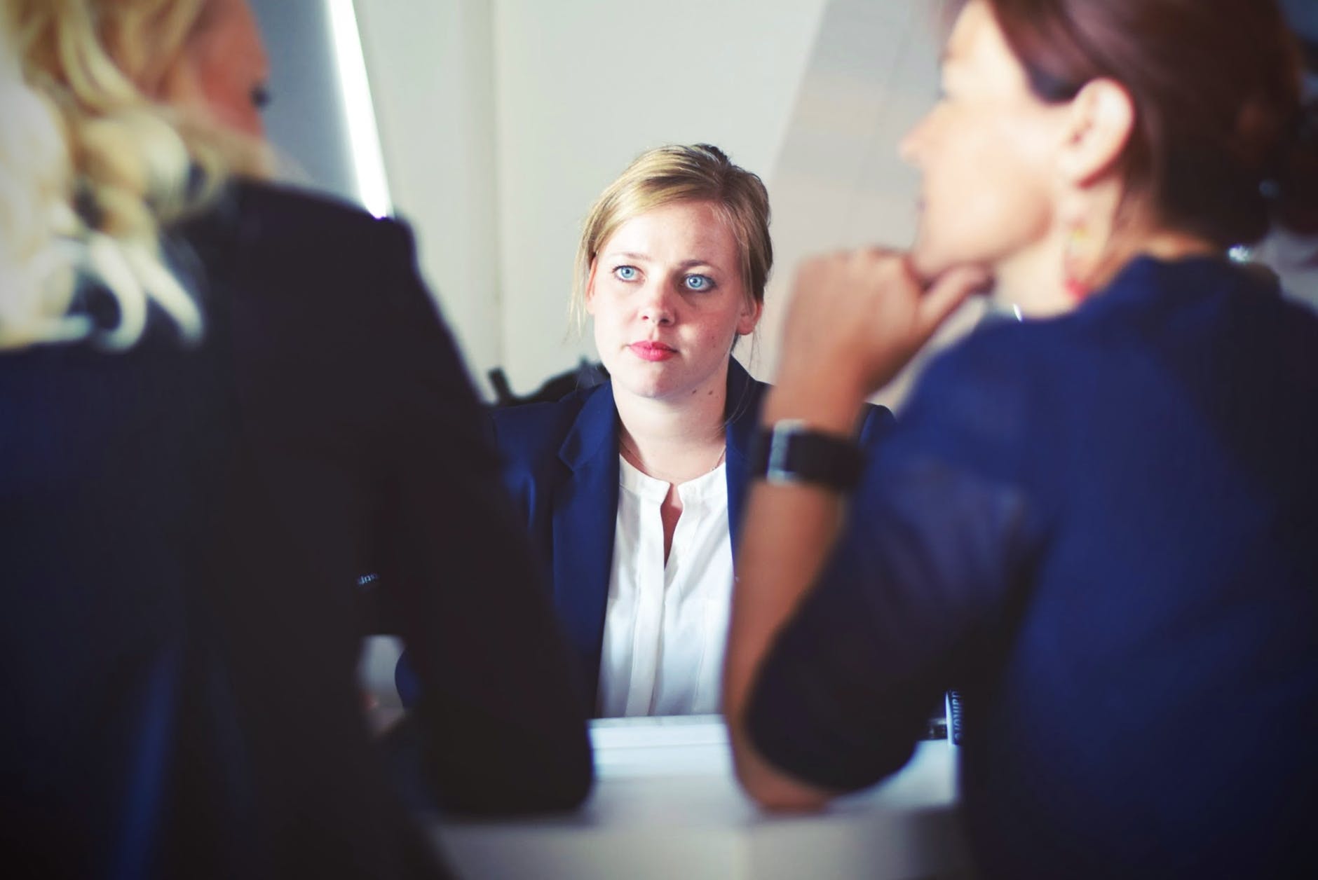 A businesswoman in a suit sitting at a desk, discussing something with her coworkers in a conference room setting.