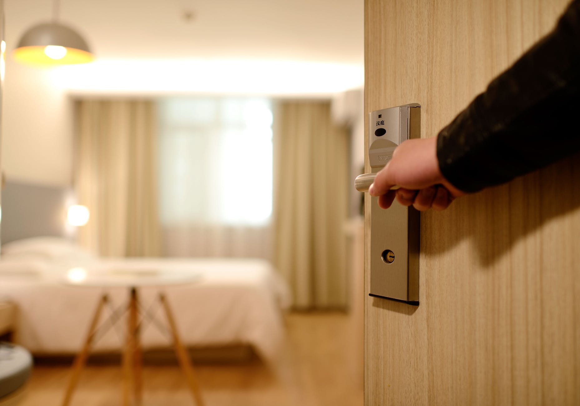 Close-up of a hand opening a hotel room door, revealing a cozy hotel room with a bed and window in the background