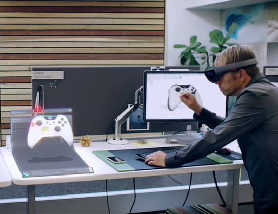 A man wearing virtual reality headset playing a video game on a large screen, with a gaming controller visible on the desk in front of him.