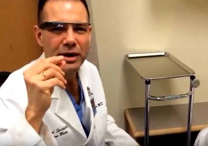 A doctor wearing a white lab coat and glasses, sitting in an office with a medical equipment in the background.