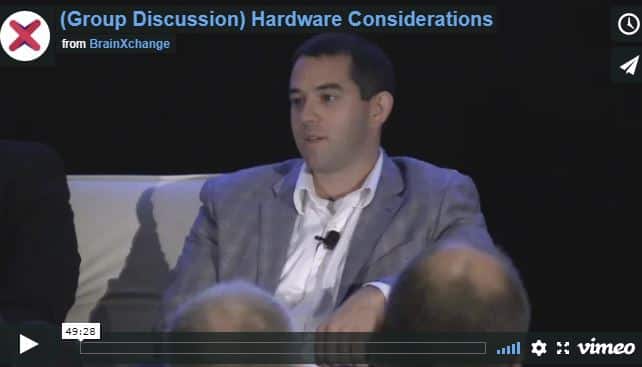 A man in a gray suit speaking at a podium with the text 'Group Discussion) Hardware Considerations' on the screen behind him