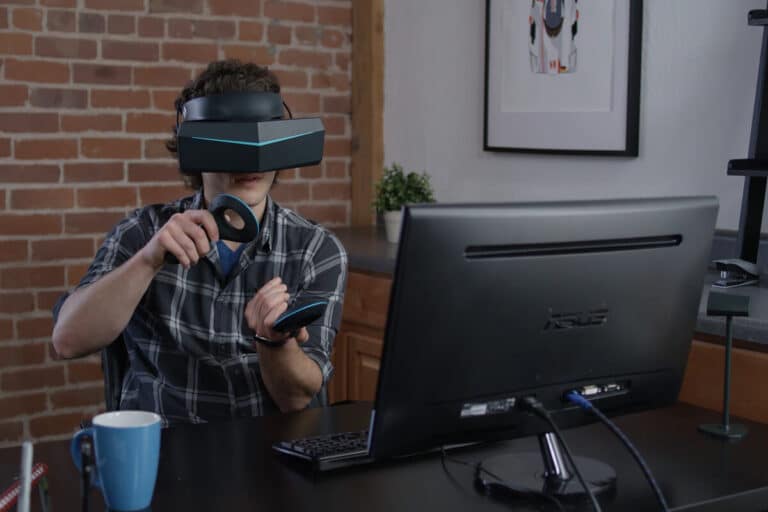 Man wearing virtual reality headset and using controllers, seated at a desk with a computer monitor in a home office setting.