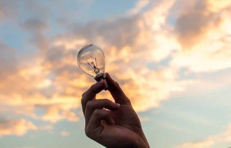 Hand holding a light bulb against a colorful cloudy sky at sunset, representing creativity and innovation