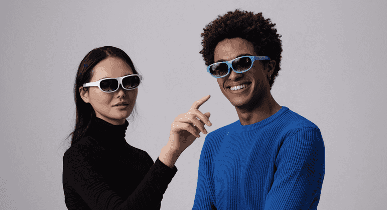 Two young people wearing sunglasses, one in a blue sweater and the other in a black turtleneck, looking at the camera and smiling