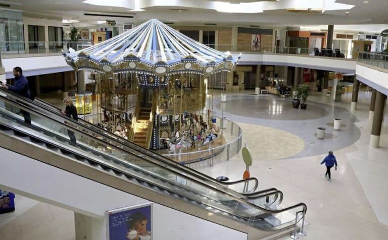 An indoor shopping mall with a large, illuminated carousel in the center, surrounded by stores and escalators