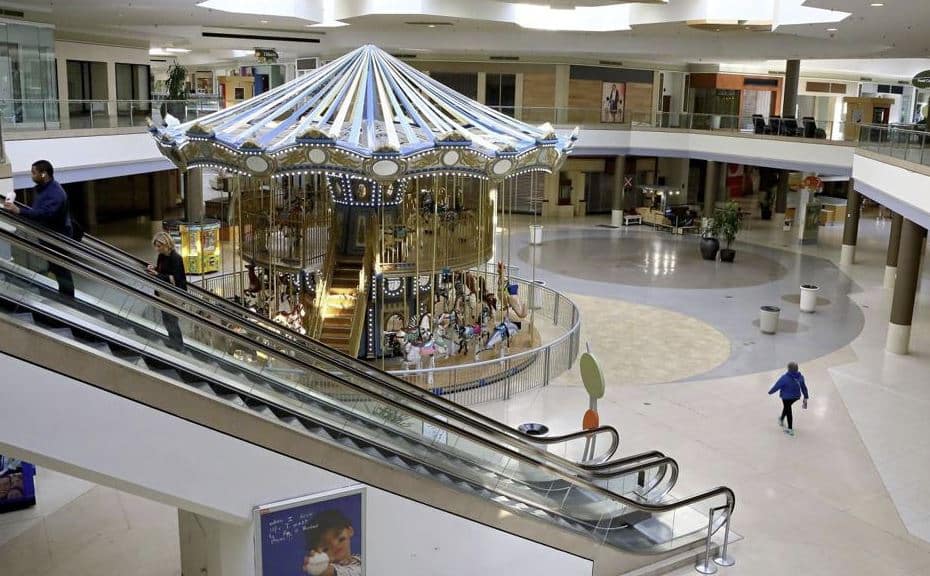 An indoor shopping mall with a large, illuminated carousel in the center, surrounded by stores and escalators