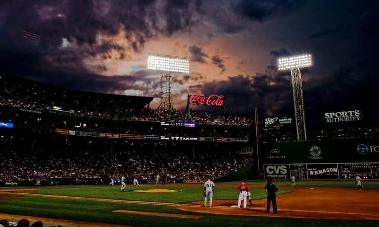 Nighttime baseball stadium with players on the field, large scoreboard and lights, and dramatic cloudy sky in the background