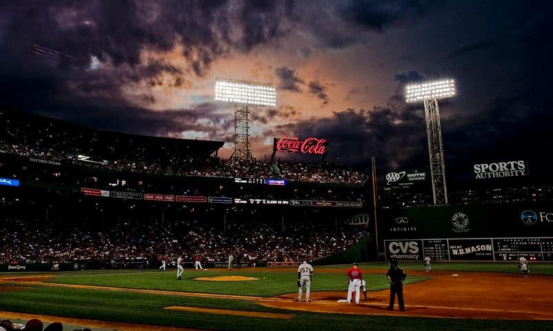 Nighttime baseball stadium with players on the field, large scoreboard and lights, and dramatic cloudy sky in the background
