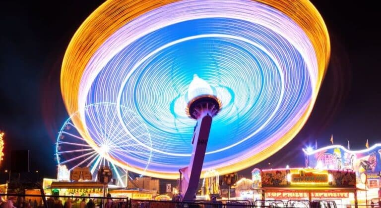Colorful, swirling lights and a large rotating ferris wheel at a fairground or carnival at night