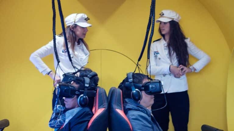 Two women in white uniforms stand in front of two people wearing virtual reality headsets, using VR technology for entertainment or training