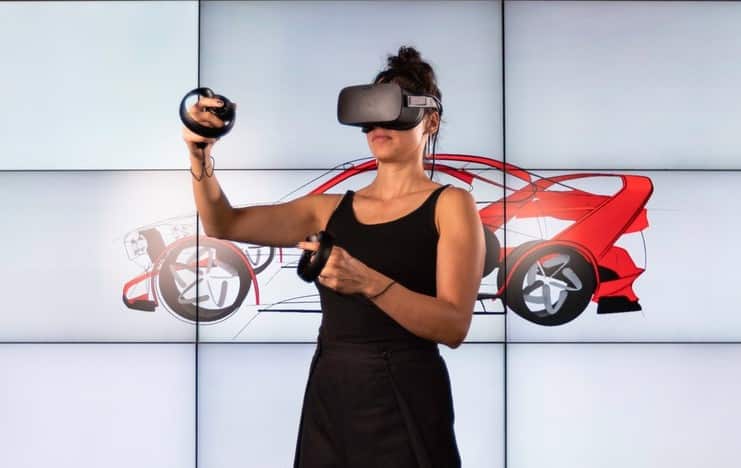 Woman wearing virtual reality headset and controllers, interacting with a digital car design on the background screen