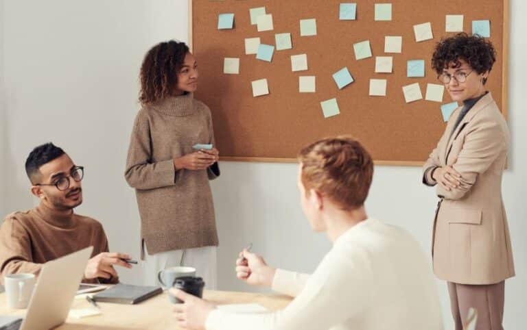 A group of four coworkers collaborating in a modern office setting, with a corkboard covered in sticky notes in the background.
