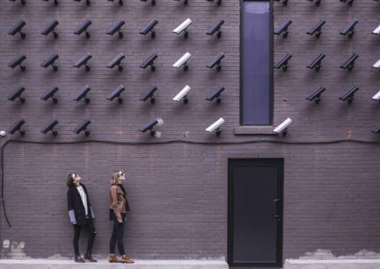 Image of a brick wall with numerous security cameras installed, with two people standing in front of the wall