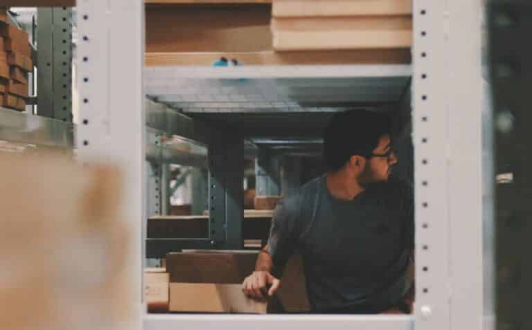 A worker wearing a gray shirt and glasses is shown organizing boxes on shelves in a warehouse