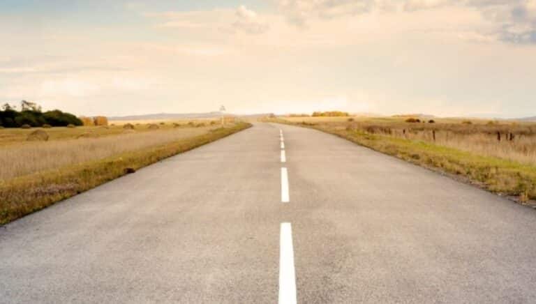 Long, straight rural road stretching into the distance through a grassy, rolling landscape with hills in the background
