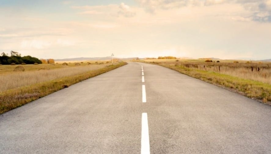 Long, straight rural road stretching into the distance through a grassy, rolling landscape with hills in the background