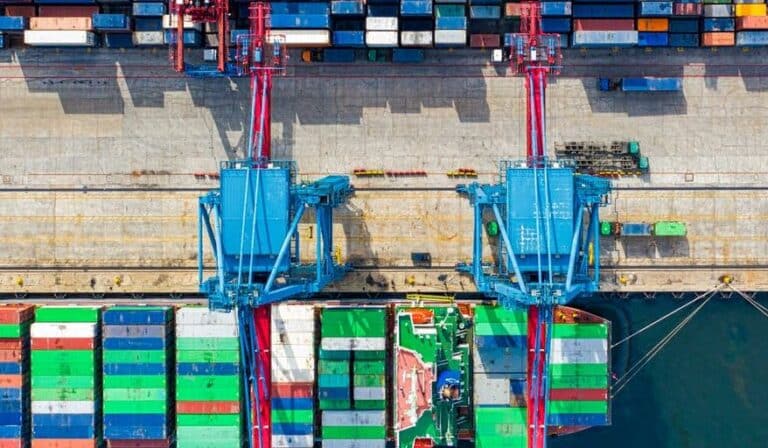 Colorful shipping containers stacked in a cargo port with two large blue cranes lifting containers