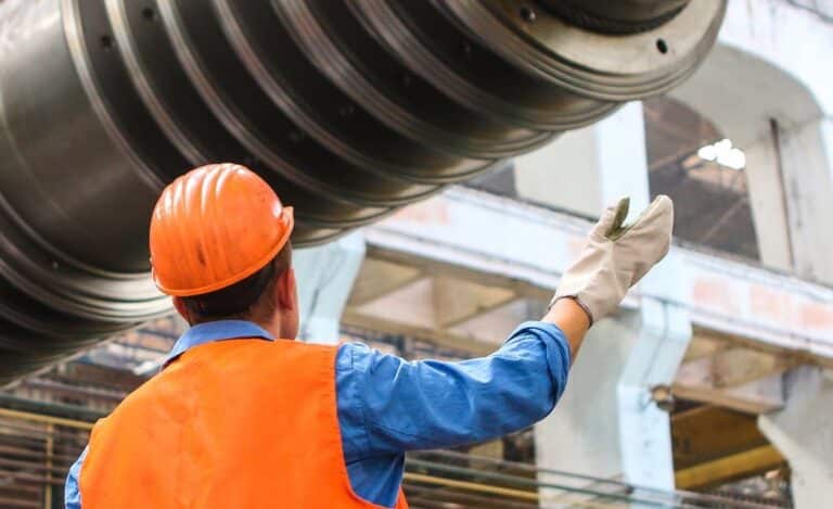 An industrial worker in a orange hard hat and blue overall inspecting machinery in a factory