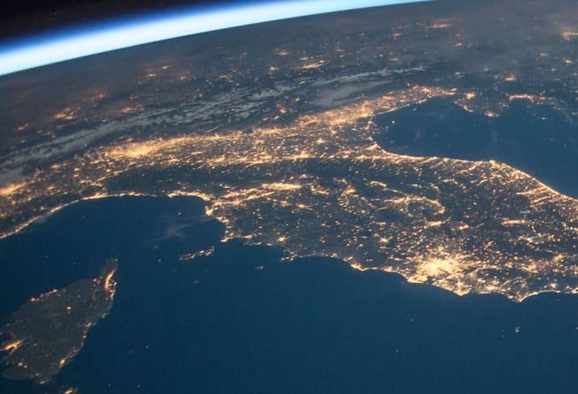 Aerial view of a city at night, with bright lights illuminating the landscape against the dark backdrop of the ocean and night sky.