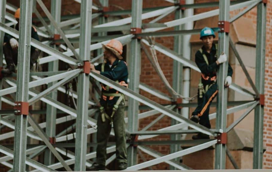 Construction workers on a metal scaffolding structure, wearing safety gear like hard hats and harnesses.