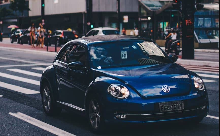 A blue Volkswagen Beetle car parked on a city street, with pedestrians in the background