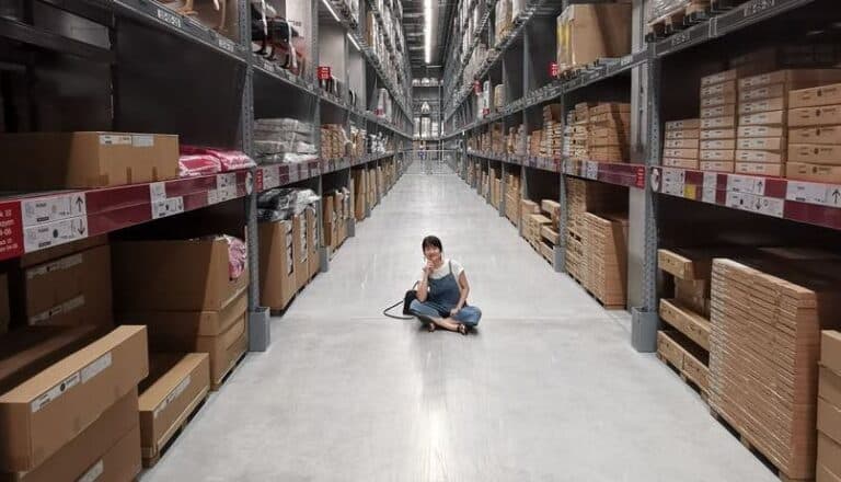 A person sitting on the floor of a large warehouse-style storage facility, surrounded by rows of shelves and boxes
