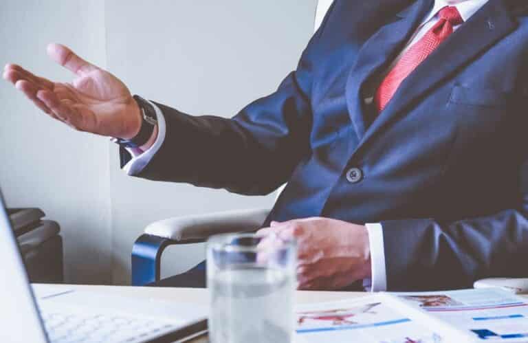 A businessman in a suit sits at a desk, gesturing with his hands while working on a laptop computer.