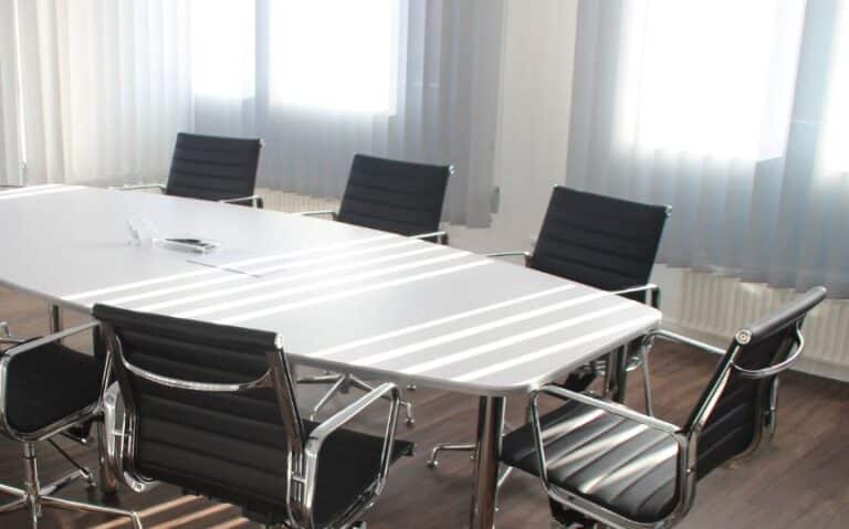 A modern conference room with a long white table and black leather chairs, surrounded by large windows