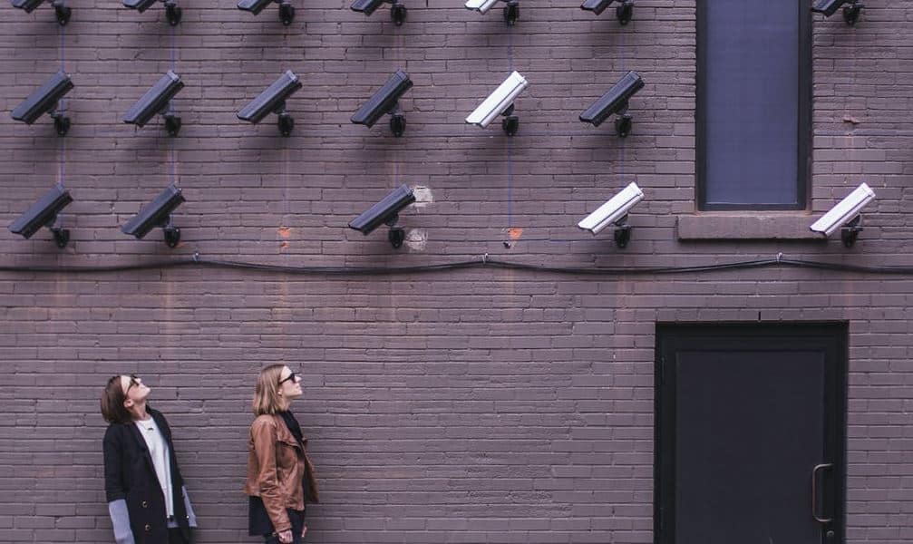 Two people walking in front of a brick wall with multiple security cameras mounted on it