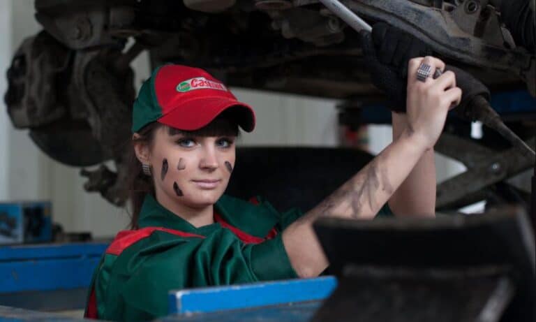 A female auto mechanic in a green uniform and red hat working on a car engine in a garage.