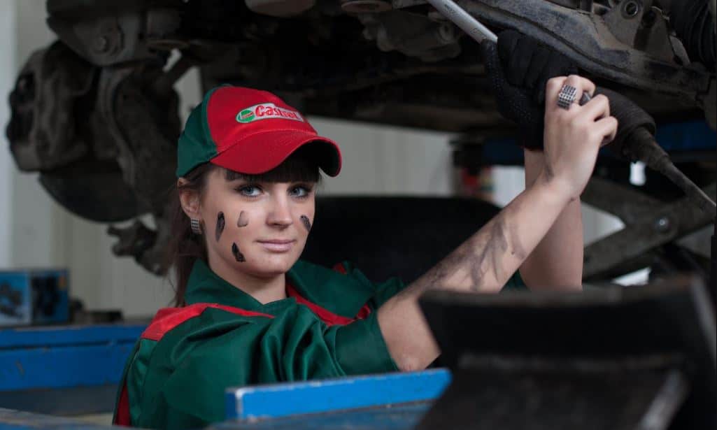 A female auto mechanic in a green uniform and red hat working on a car engine in a garage.