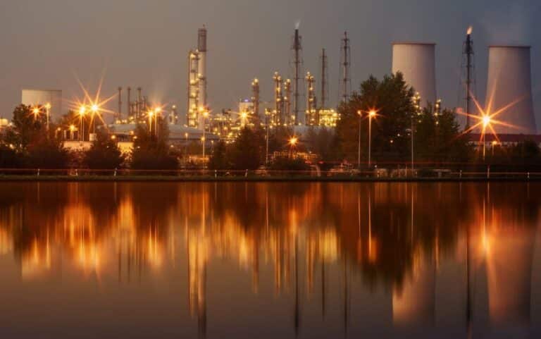 Industrial refinery plant with tall towers and tanks reflected in calm water at night, illuminated by bright lights