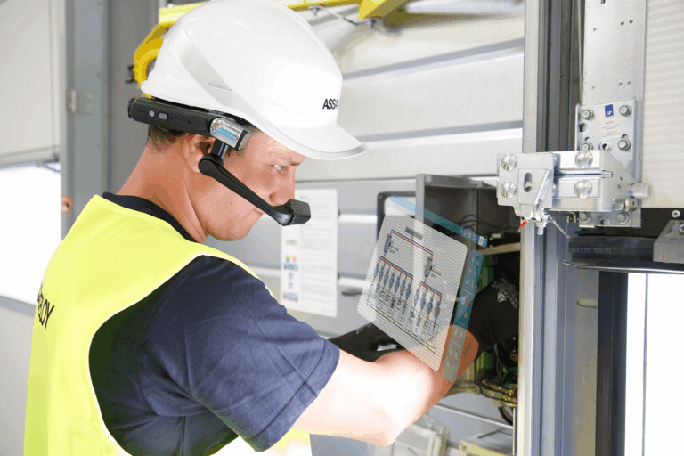 An electrician in a hard hat and safety vest working on an electrical panel with a digital tablet in his hands.