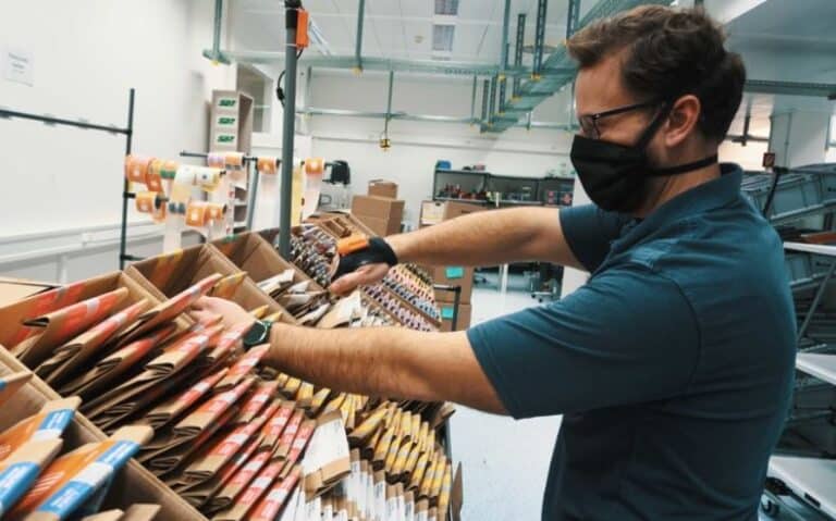 A man wearing a face mask and blue shirt is organizing and restocking shelves in a warehouse or stockroom setting.