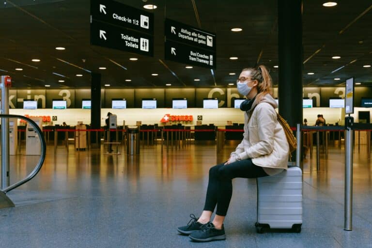 Woman sitting on luggage in an airport terminal, wearing a mask and surrounded by signs directing travelers to various gates and services