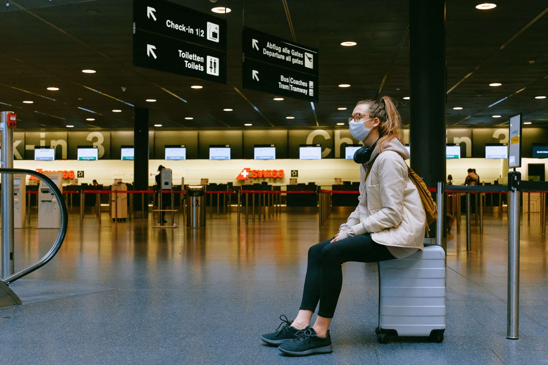 Woman sitting on luggage in an airport terminal, wearing a mask and surrounded by signs directing travelers to various gates and services